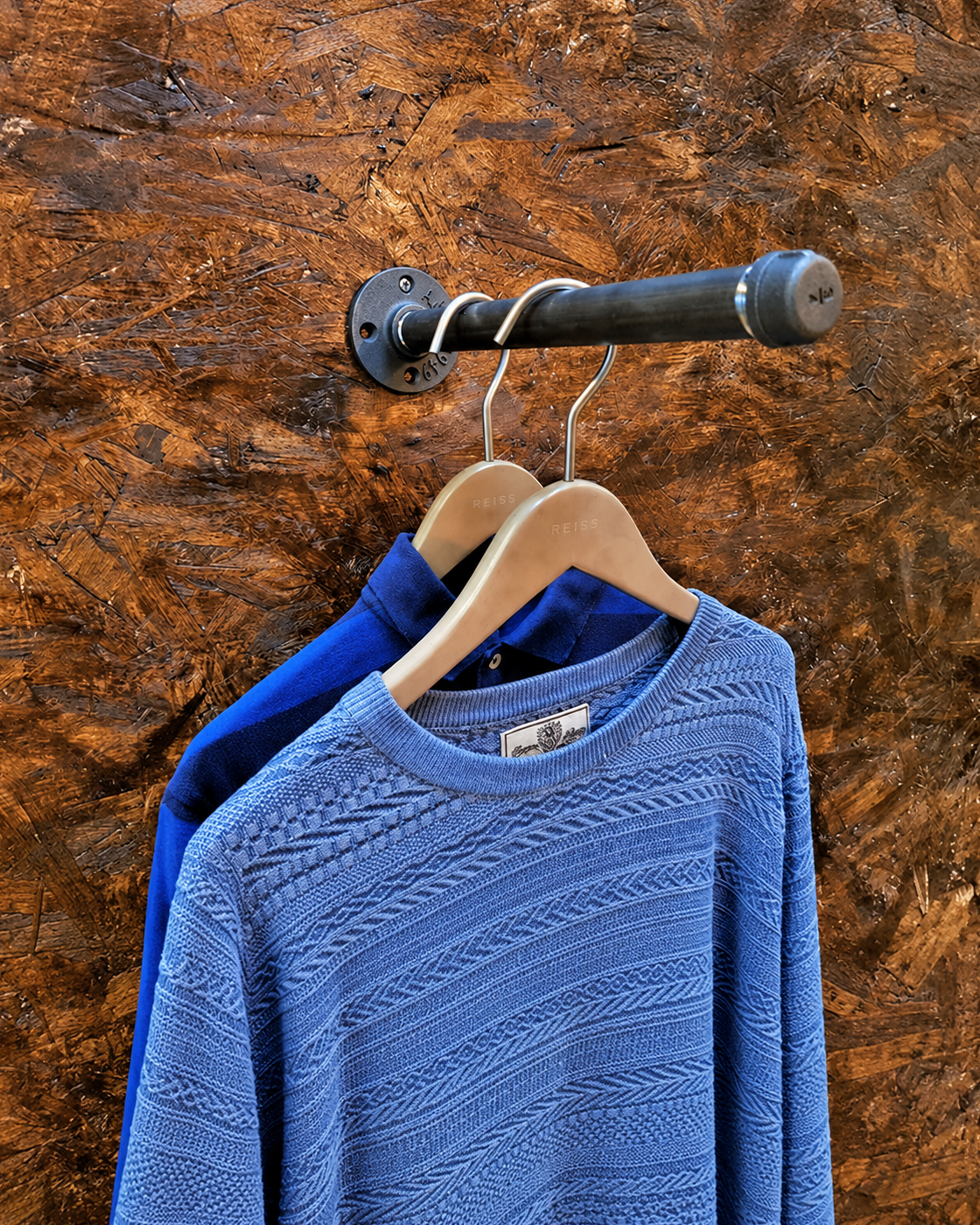 Black wall-mounted Flux pipe clothes rail holding a blue jumper and shirt against a textured wood wall.